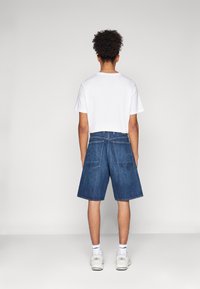 Joven con cabello rizado de pie de espaldas, vistiendo una camiseta blanca, pantalones cortos de mezclilla azul largos, calcetines blancos y zapatillas blancas.