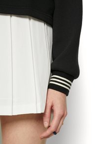 Černá mikina s pruhovanými manžetami, doplněná bílou skládací sukní. Důraz je kladen na texturovanou tkaninu a kontrastní barvy.