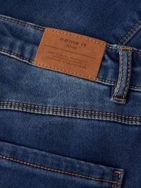 Jeans tkanina v srednje modri barvi z kontrastnimi oranžnimi šivi. Vsebuje rjavo usnjeno etiketo z znamko in podatki o negi.