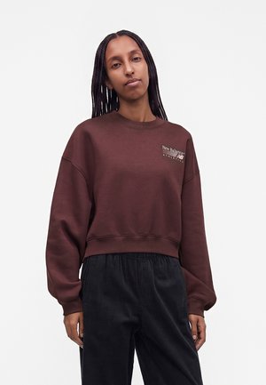 Femme aux longs cheveux tressés portant un sweat New Balance marron foncé et un pantalon noir sur un fond clair uni.