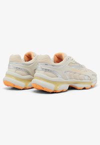 Zapatillas deportivas beige con materiales de malla y sintéticos, acentos plateados y suelas naranjas. Diseño redondeado con cuellos acolchados para mayor comodidad.