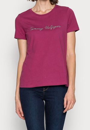 Woman wearing a short-sleeve, magenta Tommy Hilfiger t-shirt paired with dark blue jeans against a plain white background.