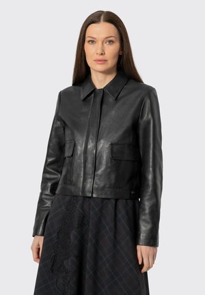 Donna con capelli castani lisci che indossa una giacca di pelle nera e una gonna a quadri con dettagli in pizzo su uno sfondo semplice.