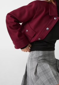 Personne portant une veste bordeaux, un pull noir et une jupe plissée à carreaux gris, posant avec une main sur la hanche sur un fond blanc.