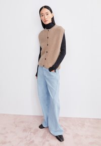 Beige gebreid vest met knopen over een zwarte coltrui, gecombineerd met lichtblauwe wijde jeans en zwarte gespen mules.