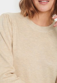 Beige stickad tröja med ribbad halsringning, texturerad tyg och puffärmsdetalj. Mjuk, lätt material med ett subtilt mönster.