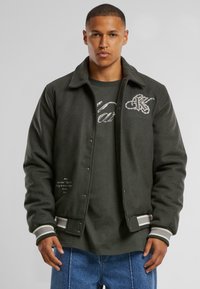 Gray wool jacket with snap buttons, ribbed cuffs, and a white embroidered "K" logo. Paired with a dark green T-shirt and blue denim.