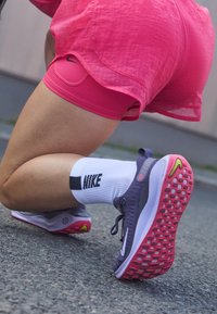 Fioletowo-białe buty do biegania Nike z teksturowaną podeszwą i różowymi akcentami, w połączeniu z białymi skarpetkami z czarnym logo, umieszczone na szarej powierzchni.