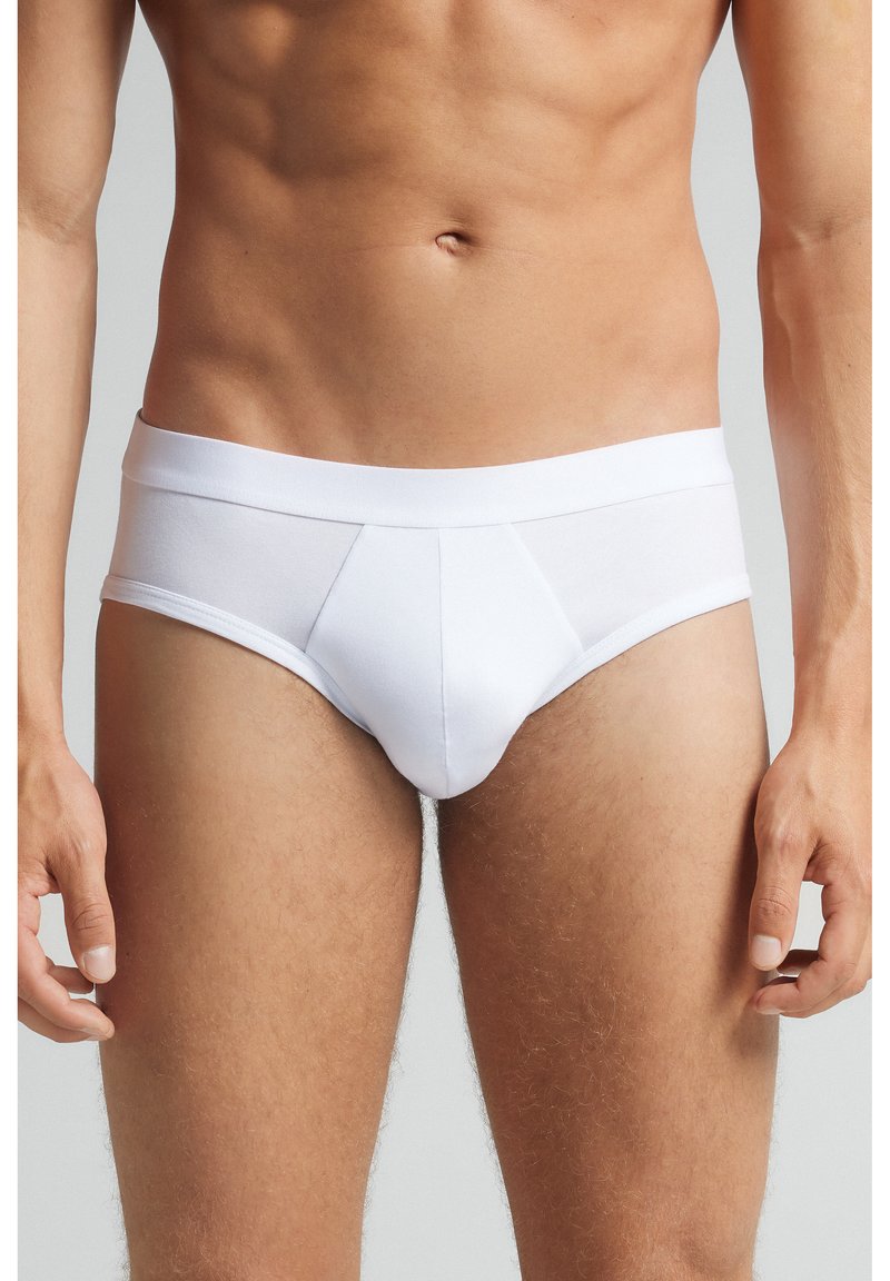 Männlicher Oberkörper in weißen Slips, zeigt einen trainierten Bauch und obere Beine, mit entspannten Händen an den Seiten vor einem hellgrauen Hintergrund.