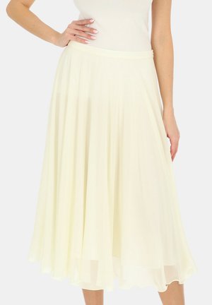 Kobieta w kremowej, bezrękawowej bluzce i zwiewnej spódnicy midi z delikatnymi plisami, stojąca z jedną ręką na biodrze.