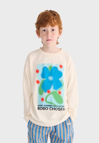 Red-haired child wearing a cream long-sleeve shirt with a large blue flower graphic and text, paired with blue and orange striped pants.