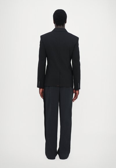 Blazer nero su misura con spalle pronunciate e chiusura a un bottone, abbinato a pantaloni neri a gamba larga. Tessuto liscio e dettagli minimi.