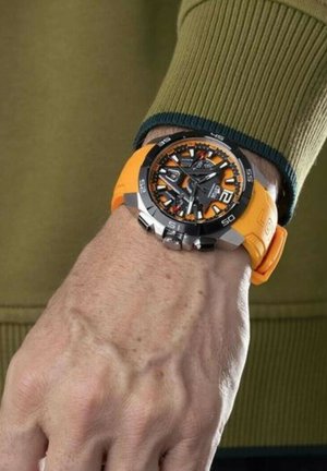 Orologio nero e arancione con quadrante rotondo, cinturino in gomma arancione, dettagli argentati e grandi numeri, indossato su una mano contro uno sfondo verde.