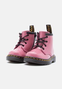 Παιδικά ροζ μπότες από γυαλιστερό δέρμα με κορδόνια, μαύρες σόλες, κίτρινη ραφή και μαύρες ταινίες με λογότυπο στο πίσω μέρος.