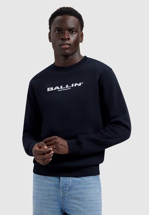 Jonge zwarte man draagt een zwart sweatshirt met het "BALLIN"-logo en lichtblauwe jeans, staand tegen een effen lichtgrijze achtergrond.