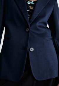 Gros plan sur une personne portant un blazer bleu marine à deux boutons sur un haut fleuri orné d'un petit nœud bleu.