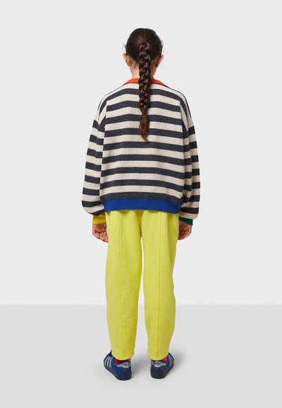 Suéter de punto a rayas en azul marino y crema con dobladillo azul, combinado con pantalones de chándal amarillos brillantes. La vista trasera muestra cabello trenzado y zapatillas.