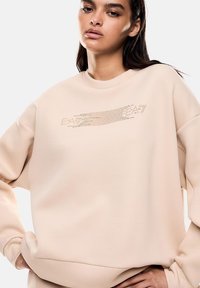 Felpa beige con scollatura rotonda e vestibilità over. Presenta un logo testurizzato sul petto in un design a contrasto. Materiale morbido.