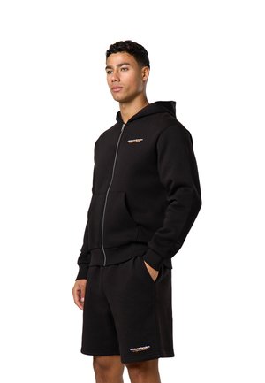 Tânăr purtând un hanorac negru cu fermoar și pantaloni scurți negri asortați, cu logo-uri mici albe și portocalii pe piept și picior.