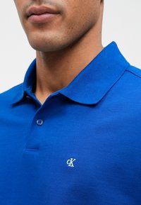 Modrá polo košeľa s textúrovanou tkaninou, s klasickým golierom a malým bielym logom "ck" na ľavej strane. Dva gombíky na zapínanie.