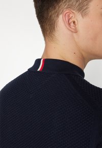 Marineblauw poloshirt met een gestructureerde stof, voorzien van een kraag met een rood-witte streepdetail aan de achterkant.