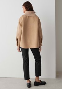 Pull en mélange de laine beige oversize avec un col côtelé haut, des épaules tombantes et des accents de boutons aux poignets, associé à un pantalon noir.