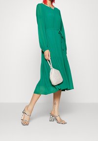 Zelené plisované šaty s dlouhými rukávy a páskem v pase, doplněné černobílými vzorovanými lodičkami a malou stříbrnou kabelkou.