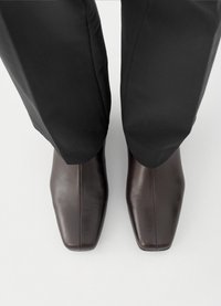 Καφέ δερμάτινα παπούτσια φόρμας με κομψό, λείο φινίρισμα και τετράγωνη μύτη. Φορεμένα με μαύρο, ραμμένο παντελόνι.