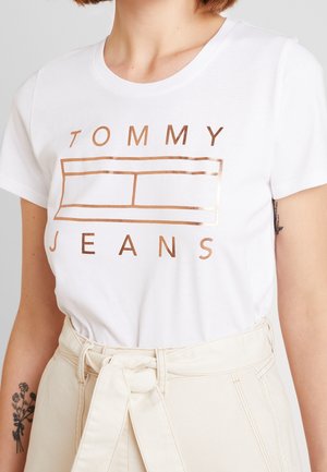 Camiseta de algodón blanca con un logo impreso en oro rosa, de manga corta, con cuello redondo y corte relajado. Combinada con pantalones beige de cintura alta.