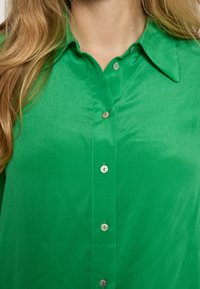 Grøn skjorte med knapper ned foran og en glat tekstur, med en spids krave og skinnende knapdetaljer. Farven er levende og mættet.