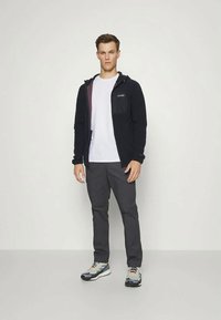 Fekete kapucnis zipzáros dzseki, szürke nadrág és színes sportcipő. A modell egyenesen áll, fehér pólót visel a dzseki alatt.