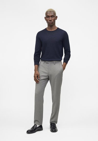 Άνδρας στέκεται με τα χέρια στις τσέπες φορώντας μπλε σκούρο μπλουζάκι μακρυμάνικο, ανοιχτό γκρι παντελόνι και μαύρα επίσημα παπούτσια σε λευκό φόντο.