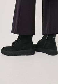 Schwarze Knöchelstiefel aus strukturiertem Leder, mit einer robusten Gummisohle, Schnürung auf der Vorderseite, Reißverschluss und Fell-Akzenten.