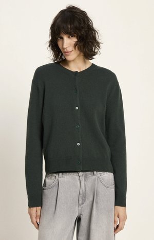 Frau mit kurzen, lockigen, dunklen Haaren, die einen dunkelgrünen, knöpfbaren Cardigan und helle, hochtaillierte Jeans trägt, vor einem schlichten Hintergrund posiert.