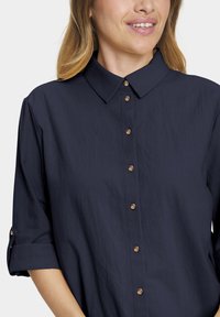 Woman wearing navy blue button-up shirt with rolled-up sleeves and golden buttons, smiling slightly.