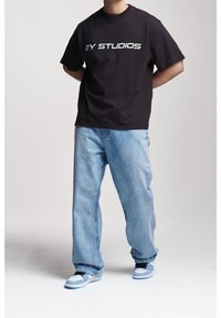 Svart bomullst-shirt med "2Y STUDIOS" tryckt i vitt, kombinerad med ljusblåa baggyjeans och sneakers med blå detaljer.