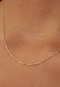 Collier en chaîne dorée reposant sur la peau, présentant de petites formes ovales liées. Texture lisse, design fin avec un éclat subtil.