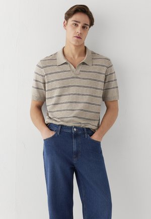 Junger Mann trägt ein beiges, gestreiftes Kurzarm-Polohemd und blaue Jeans, steht mit den Händen in den Taschen vor einer weißen Wand.