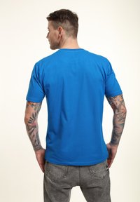 Blå kortærmet t-shirt med rund halsudskæring og glat tekstur, parret med grå jeans. Synlige tatoveringer på armene.