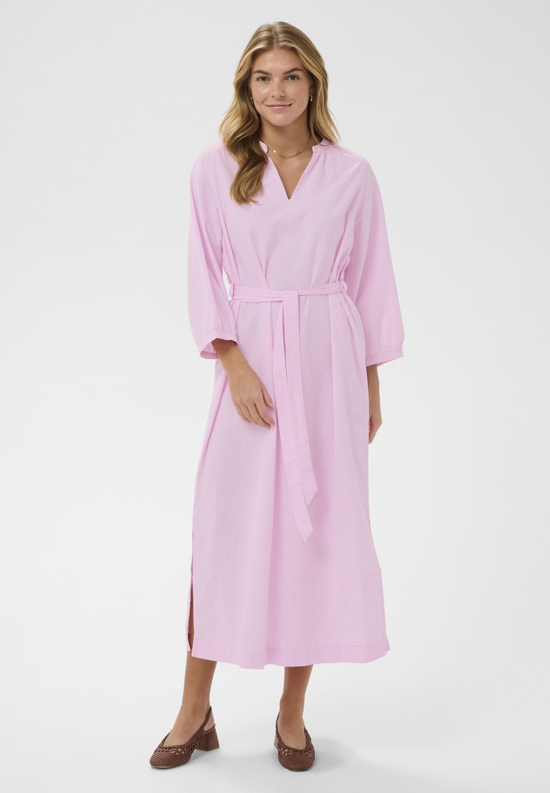 Femme aux longs cheveux blonds portant une robe midi rose clair ceinturée et des chaussures tressées marron à talons blocs, debout sur un fond blanc.