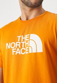Orange bomulds T-shirt med et stort hvidt logo med teksten "THE NORTH FACE." Rund hals og korte ærmer. Glat tekstur.