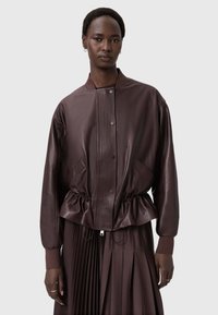 Hnědá kožená bomber bunda s ribovanou strukturou, patentkami, bočními kapsami a shromážděným pasem. Nosená přes plisovanou hnědou sukni.