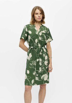 Femme portant une robe verte longueur genou avec manches courtes, motifs floraux blancs et ceinture nouée à la taille, debout devant un fond blanc uni.