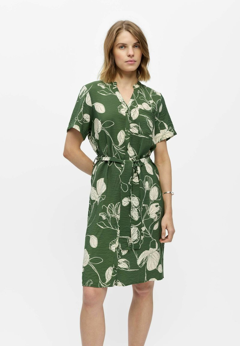 Femme portant une robe verte longueur genou avec manches courtes, motifs floraux blancs et ceinture nouée à la taille, debout devant un fond blanc uni.