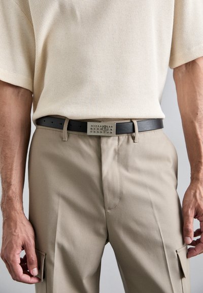 Czarny skórzany pasek z prostokątną metalową klamrą z wytłoczonymi kropkami i cyframi, noszony z beżowymi spodniami i teksturowaną kremową bluzką.