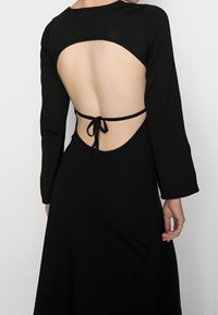 Abito lungo nero a maniche lunghe con un design a schiena scoperta e chiusura con lacci. Tessuto dalla texture liscia e silhouette fluida.