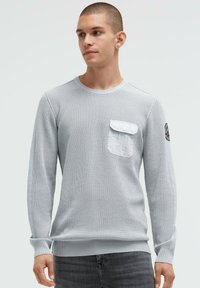 Graues Langarmshirt mit strukturiertem Strickdesign, einer Brusttasche und gerippten Bündchen. Der Stoff wirkt weich und hat eine lockere Passform.