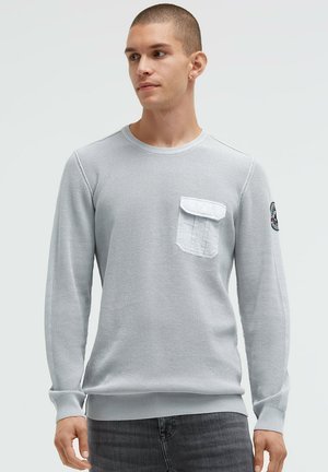 Graues Langarmshirt mit strukturiertem Strickdesign, einer Brusttasche und gerippten Bündchen. Der Stoff wirkt weich und hat eine lockere Passform.