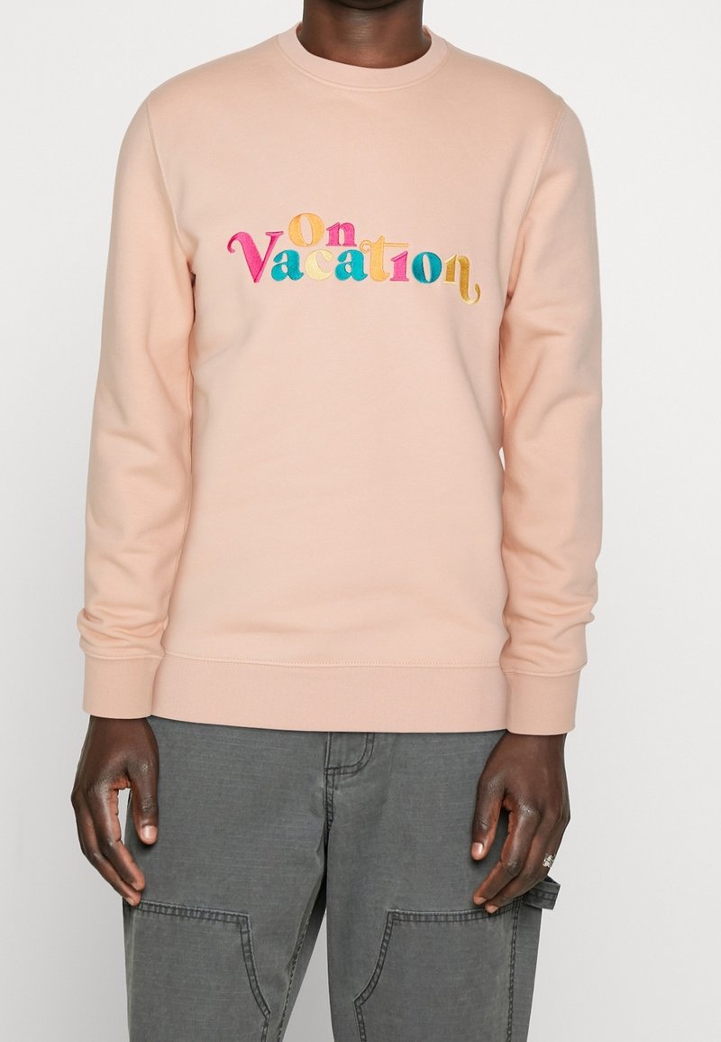 Sweat-shirt pêche avec un texte multicolore brodé indiquant "On Vacation". Comprend un col ras du cou ainsi que des poignets et un ourlet côtelés.