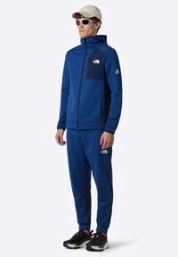 Blauwe sportieve trainingspak met een met rits afgesloten jas en broek. Kenmerkt zich door een borstzak, contrasterende zwarte accenten en een rubberen logo. Geweven stof.